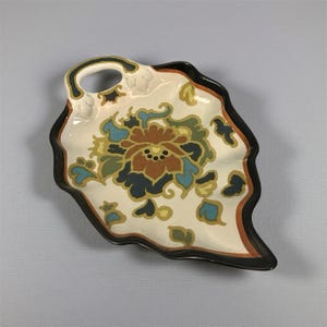 May include: A leaf-shaped ceramic serving dish with a handle. The dish has a cream base with a floral design in brown, blue, and green. The rim is black with an orange border. A decorative piece for serving snacks, or small items.