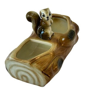 May include: A ceramic planter shaped like a tree stump, featuring a small squirrel figurine perched on top. The planter has two rectangular openings and is painted in shades of brown and cream, with a glossy finish. The squirrel is light brown with a dark brown tail.