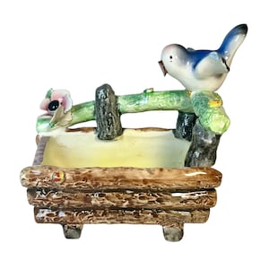 May include: A ceramic bird ornament perched on a miniature wooden fence. The bird has blue and white plumage, with a brown beak. The fence is painted green and brown, and the base is a light yellow colour. A pink flower is on the left side of the fence.