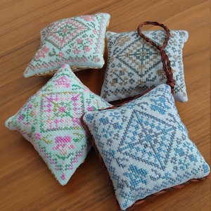 Puede incluir: Cuatro pequeñas almohadas cuadradas bordadas con intrincados patrones geométricos y florales en tonos rosa, verde, azul y marrón. Cada almohada tiene un diseño y una combinación de colores diferentes. Una tiene un lazo y una borla trenzados marrones.