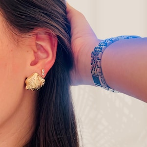 Pode incluir: Brincos dourados em forma de concha com detalhes intrincados. Um pequeno brinco de pressão transparente também é visível. Um relógio prateado com pulseira de elos está no pulso.