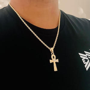 May include: A gold chain necklace with a gold ankh pendant. The ankh has intricate details and a small white stone in the centre.
