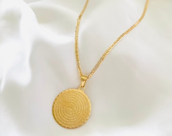 May include: A gold chain necklace with a round pendant featuring a circular inscription in Spanish. The pendant has a textured edge.
