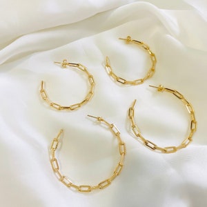 May include: Four gold-colored hoop earrings with a chain link design. The earrings are arranged on a white, silky fabric. The earrings are a simple, yet elegant accessory.