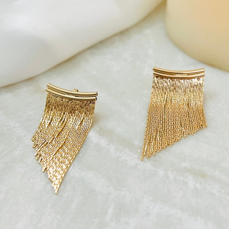 May include: A pair of gold dangle earrings with a fringe design. The earrings are made of a chain-like material and have a curved top.