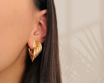 May include: Gold-toned, textured, geometric hoop earrings. The earrings are worn by a person with long brown hair.