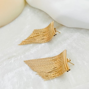 May include: A pair of gold-toned dangle earrings with a fringe design. The earrings are made of a thin, chain-like material and have a curved bar at the top.