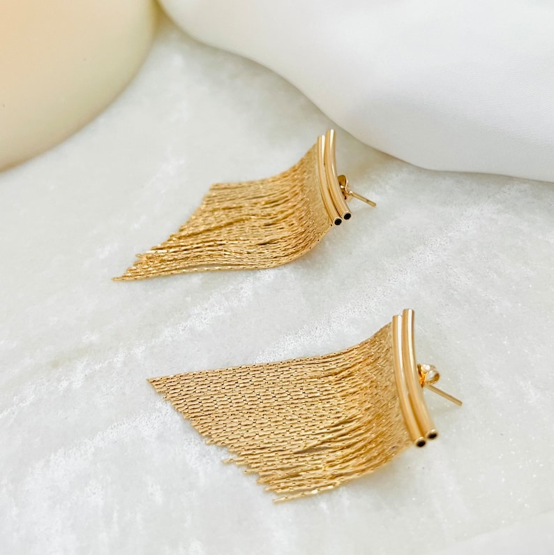 May include: A pair of gold-toned dangle earrings with a fringe design. The earrings are made of a thin, chain-like material and have a curved bar at the top.