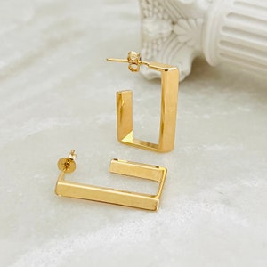 May include: A pair of gold-toned square hoop earrings. The earrings are made of thick metal and have a simple, modern design.