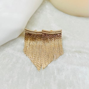 May include: A pair of gold-toned dangle earrings with a fringe design. The earrings are made of a chain-like material and have a curved bar at the top.