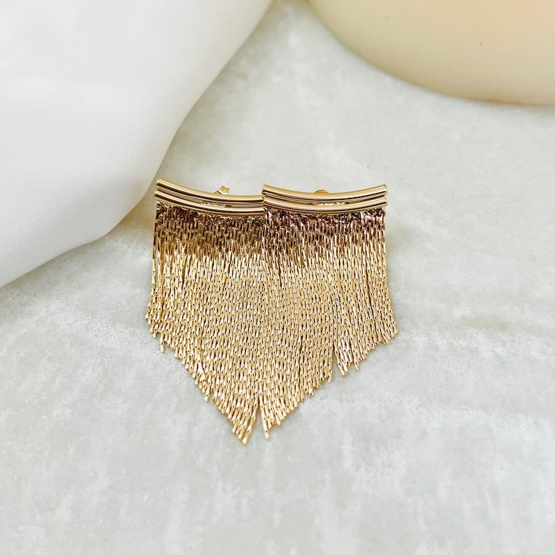 May include: A pair of gold-toned dangle earrings with a fringe design. The earrings are made of a chain-like material and have a curved bar at the top.
