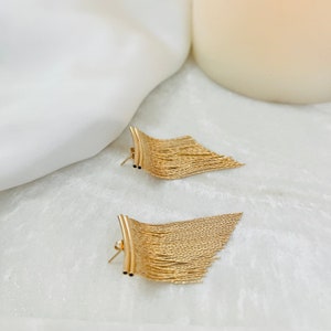 May include: A pair of gold-toned earrings with a fringe design. The earrings are made of a thin, chain-like material and have a curved, bar-like top.