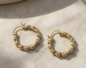 May include: A pair of gold hoop earrings featuring a textured bamboo-inspired design. Each circular earring has a hinged post closure and a polished metallic finish that reflects light, creating a detailed, segmented appearance across the surface.