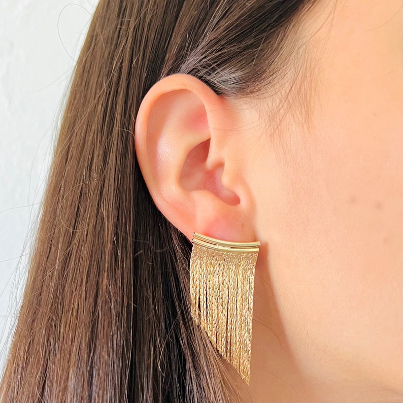 May include: Gold dangle earrings with a fringe design. The earrings are made of a chain-like material and have a curved top.