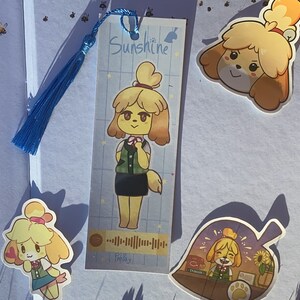 May include: A set of three bookmarks featuring a cartoon dog character with yellow fur, brown eyes, and a pink nose. The bookmarks are printed with a light blue background and have a white border. The text "Sunshine" is printed on one bookmark. The text "Play!" is printed on another bookmark. The third bookmark features the dog character standing behind a counter with a leaf-shaped sign that says "Nook's Cranny".