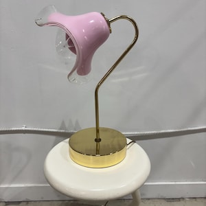 May include: A vintage table lamp featuring a pink, flower-shaped glass shade with a clear glass interior. The lamp has a brass-coloured, curved stem and a round base. The lamp is on a white stool.