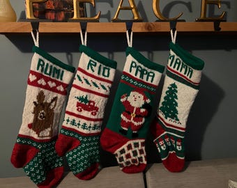 Könnte beinhalten: Vier gestrickte Weihnachtsstrümpfe hängen in einer Reihe. Der erste zeigt ein Rentier und den Namen RUMI. Der zweite zeigt einen roten Lastwagen und den Namen RIO. Der dritte zeigt den Weihnachtsmann und den Namen PAPA. Der vierte zeigt einen Baum und den Namen MAMA.
