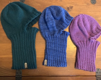 May include: Three knitted balaclavas in teal, blue, and purple. Each balaclava has a ribbed texture and a small tag attached to the side.