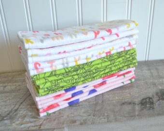 May include: A stack of colorful, patterned baby washcloths. The washcloths feature various designs, including letters, floral patterns, and other playful graphics. The colors include pink, green, yellow, and blue. The washcloths are stacked neatly on a wooden surface.