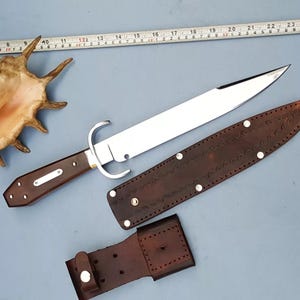 May include: A large, polished steel knife with a brown wooden handle and a curved guard. The knife is accompanied by a brown leather sheath and a belt loop. A seashell and a measuring tape are also present.