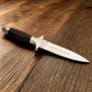 Puede incluir: Una daga de plata con mango negro. La hoja es afilada y puntiaguda. El mango es de madera y tiene un acabado liso.