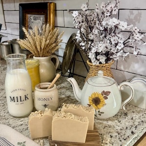 May include: A kitchen still life with a milk bottle, honey pot, and three bars of soap. The milk bottle has "Farm Fresh Milk" text. A teapot with a sunflower design and a vase of white flowers are also present.