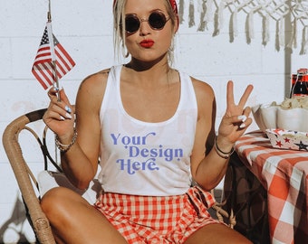 Könnte beinhalten: Eine Frau trägt ein weißes Tanktop mit dem Text "Your Design Here" in Lila, rot-weiß karierte Shorts und weiße Sandalen. Sie hält eine kleine amerikanische Flagge und lächelt. Sie sitzt auf einem Korbstuhl vor einer weißen Backsteinwand.