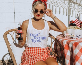 Könnte beinhalten: Eine Frau trägt ein weißes Tanktop mit dem Text "Your Design Here" in Lila, einen rot-weiß karierten Rock und ein rotes Bandana-Stirnband. Sie sitzt auf einem Korbstuhl und hält eine Glasflasche Coca-Cola in der Hand. Sie trägt eine Sonnenbrille und hat rote Fingernägel.
