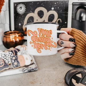 May include: A white enamel mug with a black rim and handle. The mug has a pink design that says "Your design here" with stars around the text. The mug is being held by a hand with black nail polish.