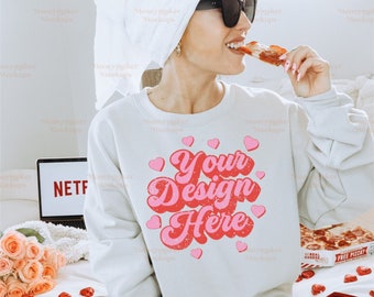 Puede incluir: Una sudadera blanca con un gráfico rosa y rojo que dice "Your Design Here" rodeado de corazones rosas. La sudadera la lleva puesta una persona sentada en una cama con un portátil, rosas y una caja de bombones.