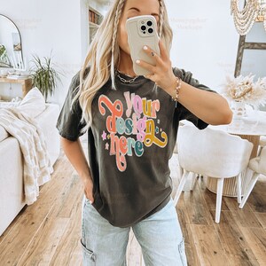 May include: A black t-shirt with the text "Your design here" in colourful letters. The shirt is being worn by a person with long blonde hair and blue jeans.