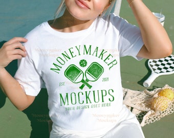 May include: A white t-shirt with a green circle graphic featuring the text "MONEYMAKER" and two crossed paddleball rackets. The text "EST. 2021" and "MOCKUPS YOUR DESIGN GOES HERE" are also included in the graphic.