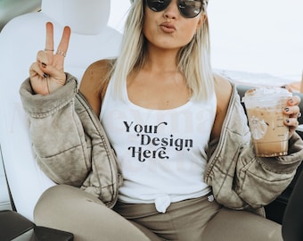 May include: A woman wearing a white tank top with the text "Your Design Here" in black lettering, a black baseball cap with a peace sign, and sunglasses. She is sitting in a car and holding a cup of iced coffee.