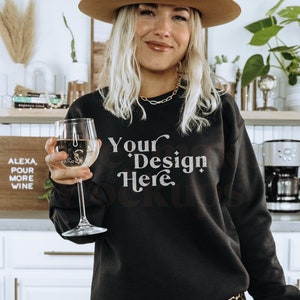 May include: A woman wearing a black sweatshirt with the text "Your Design Here" in white lettering. She is holding a glass of white wine and wearing a brown hat and leopard print pants.
