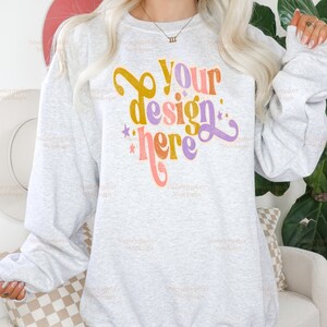 Pu&ograve; includere: Una felpa grigio chiaro con la scritta "Your design here" in un font retr&ograve;. Il testo &egrave; nei toni dell'oro, del viola, dell'arancione e del rosa. La felpa ha maniche lunghe e girocollo.