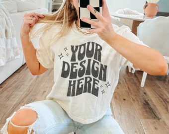 May include: A woman wearing a white t-shirt with the text "YOUR DESIGN HERE" in a retro font. She is wearing light blue jeans with distressed details and a black baseball cap.