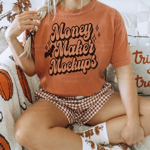 May include: A woman wearing a burnt orange t-shirt with the text "Money Maker Mockups" in a retro font. She is sitting on a white and orange blanket with a pumpkin pattern.