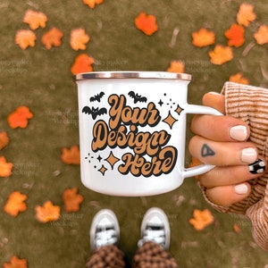 Puede incluir: Taza de camping de esmalte blanco con un dise&ntilde;o dorado y negro que dice "Your Design Here". La taza est&aacute; sostenida por una mano con un tatuaje en el dedo. El fondo es una imagen borrosa de hojas de oto&ntilde;o.
