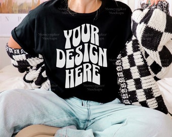 May include: A black t-shirt with the text "YOUR DESIGN HERE" in white. The shirt is being worn by a person sitting on a white bed with a black and white checkered blanket.