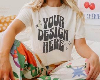 May include: A woman wearing a white t-shirt with the text "YOUR DESIGN HERE" in a retro font. The shirt is a blank canvas for customisation. The woman is sitting on a bed with a colourful patterned blanket and a book.