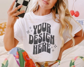 May include: White t-shirt featuring a black and white graphic that reads "YOUR DESIGN HERE". The shirt is being worn by a person sitting on a bed with a book and glasses.
