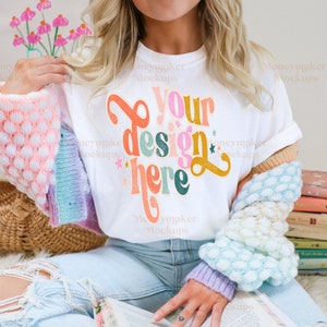 Op de afbeelding: Een wit T-shirt met de tekst "Your design here" in kleurrijke, verweerde letters. Het shirt wordt gedragen door iemand die blauwe jeans en een kleurrijke cardigan draagt.