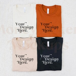 May include: Four folded t-shirts in different colours: beige, light brown, orange, and black. Each shirt has the text "Your Design Here" printed on the front.