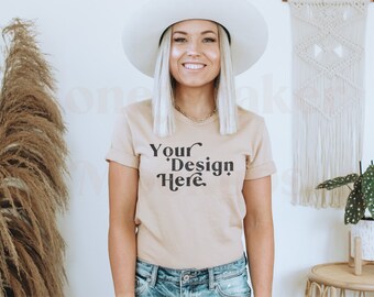 May include: A woman wearing a beige t-shirt with the text "Your Design Here" in black. She is wearing a white hat and blue jeans. The background is a white wall with a macrame wall hanging and a potted plant.