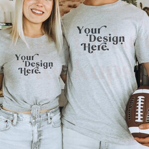 Pu&ograve; includere: Due persone indossano magliette grigie con la scritta "Your Design Here" stampata su di esse. La persona a destra tiene un pallone da football marrone.