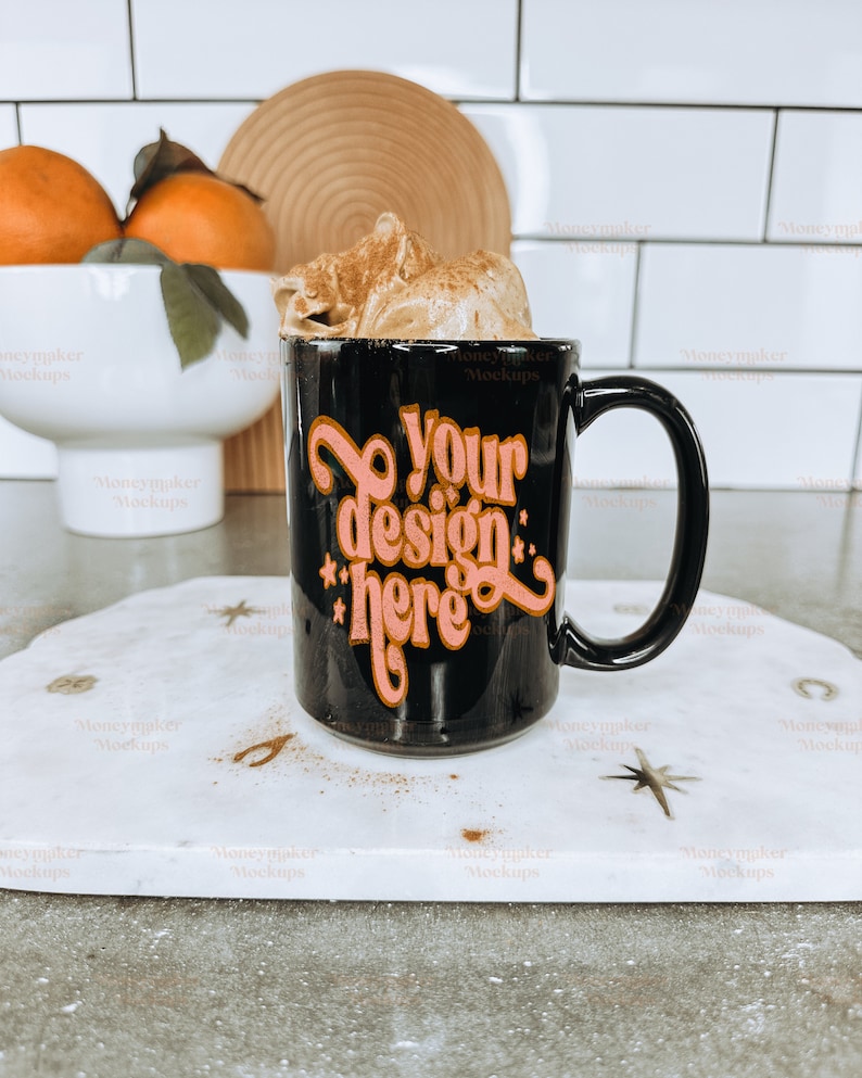 K&ouml;nnte beinhalten: Eine schwarze Kaffeetasse mit dem Text "Your design here" in Orange und Rosa. Die Tasse ist mit einem Schlagsahne-Topping gef&uuml;llt. Die Tasse steht auf einem wei&szlig;en Marmortablett mit sternf&ouml;rmigen Dekorationen.