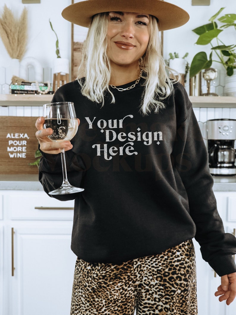 May include: A woman wearing a black sweatshirt with the text "Your Design Here" in white lettering. She is holding a glass of white wine and wearing a brown hat and leopard print pants.