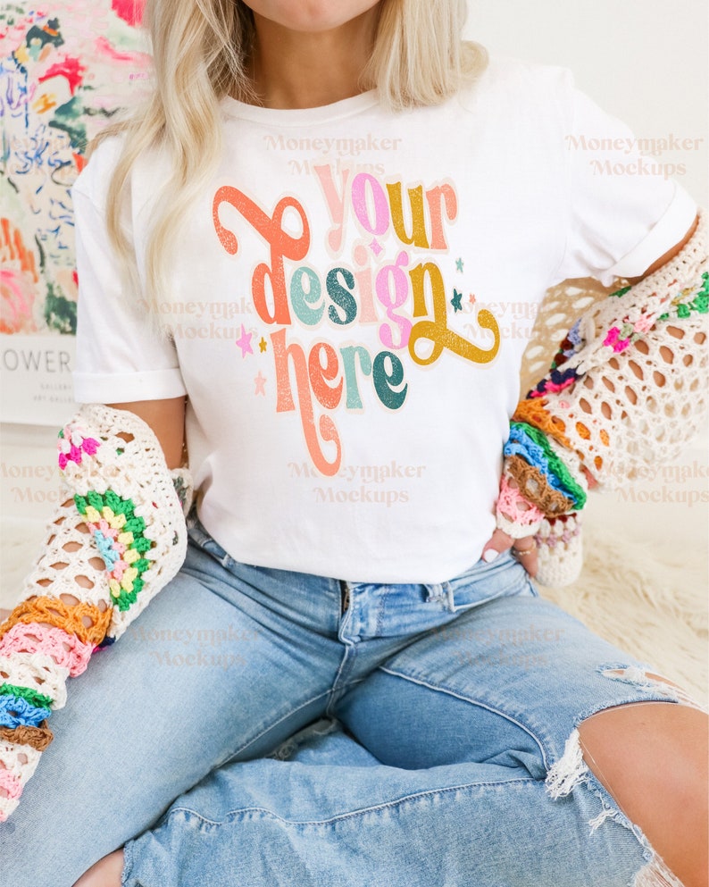 K&ouml;nnte beinhalten: Ein wei&szlig;es T-Shirt mit einer bunten Grafik, auf der "Your design here" steht. Das Shirt wird von einer Person getragen, die blaue Jeans und eine geh&auml;kelte Strickjacke tr&auml;gt.