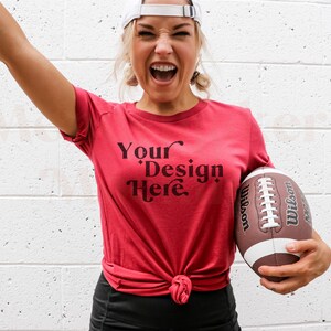 Pode incluir: Uma mulher vestindo uma camiseta vermelha com o texto "Your Design Here" em preto. Ela est&aacute; segurando uma bola de futebol marrom e branca.