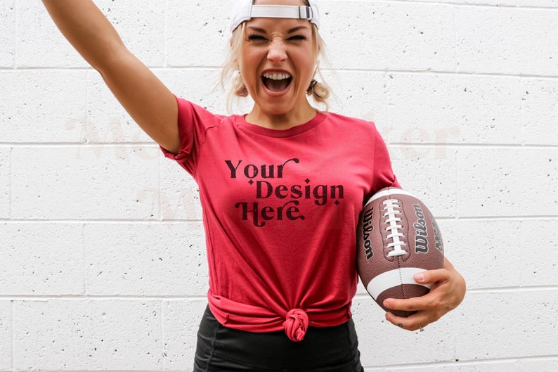 Pode incluir: Uma mulher vestindo uma camiseta vermelha com o texto "Your Design Here" em preto. Ela est&aacute; segurando uma bola de futebol marrom e branca.